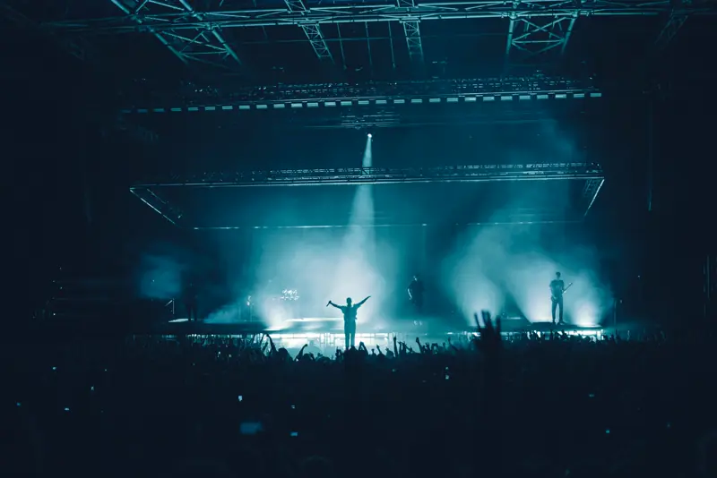 Silhouetted performer on a hazy concert stage under teal spotlights above a crowd